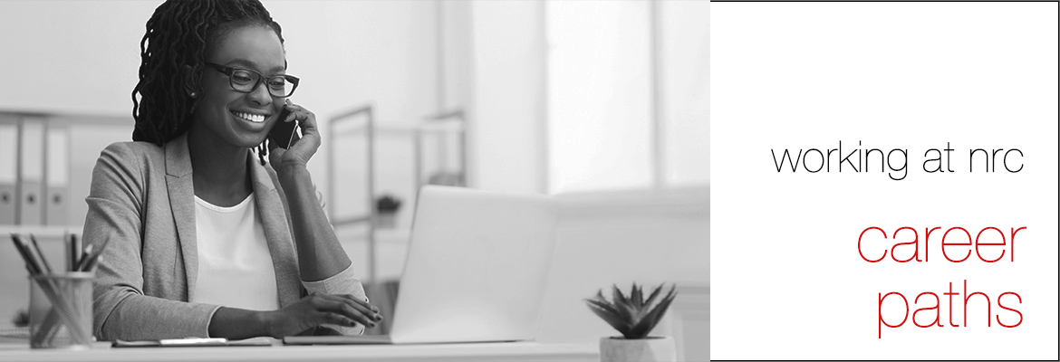 Black and white photo of a woman seated at a desk, smiling and looking at a laptop computer while talking on a cell phone.  To the right of the photo, the words 'working at nrc' and (in red font) 'career paths' on a white background.