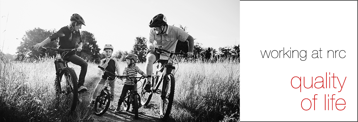 Black and white photo of a family consisting of two adults on either side of two small boys all on bicycles pausing for a moment on a bike trail in the middle of an overgrown nature field. The adults are looking down at the two small boys in the middle and all are smiling. To the right on a white background, the words 'working at nrc' and (in red font) 'quality of life'.
